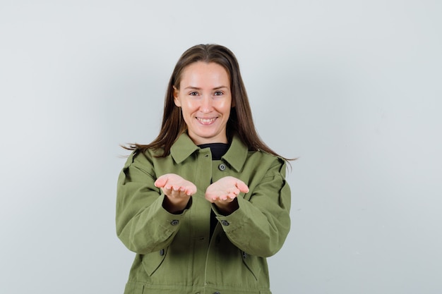 Mujer joven en chaqueta verde posando como compartir algo con alguien y mirando complacido, vista frontal.