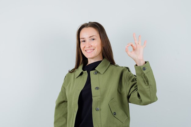 Mujer joven con chaqueta verde que muestra un gesto aceptable y una mirada optimista, vista frontal.