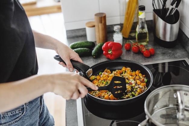 Mujer joven cocinando verduras frescas en una sartén en casa.