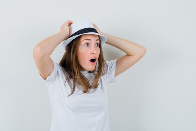 Mujer joven cogidos de la mano en la cabeza con camiseta blanca, sombrero y mirando sorprendido.