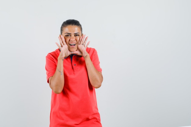 Mujer joven cogidos de la mano cerca de la boca como llamando a alguien en camiseta roja y luciendo bonita. vista frontal.