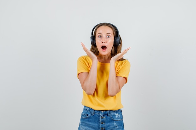 Foto gratuita mujer joven cogidos de la mano cerca de la cara en camiseta, pantalones cortos, auriculares y mirando asustado