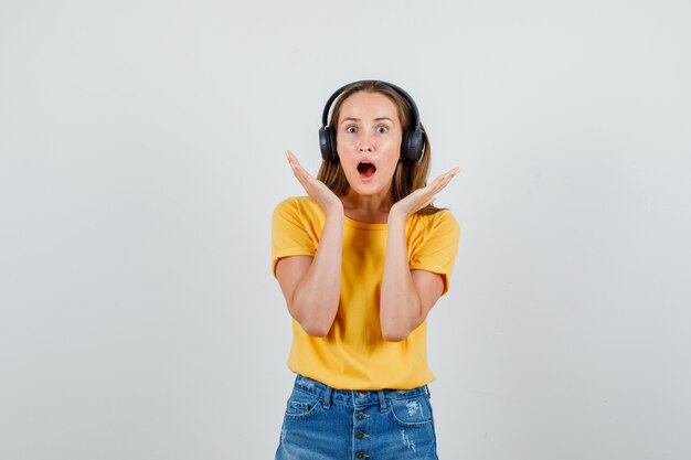 Mujer joven cogidos de la mano cerca de la cara en camiseta, pantalones cortos, auriculares y mirando asustado