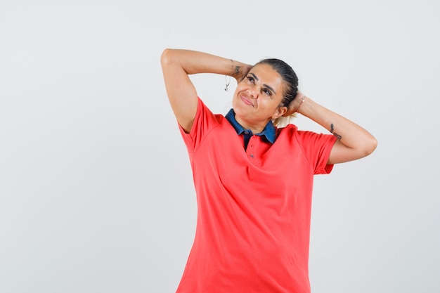 Mujer joven cogidos de la mano detrás de la cabeza, pensando en algo en camiseta roja y mirando pensativo. vista frontal.