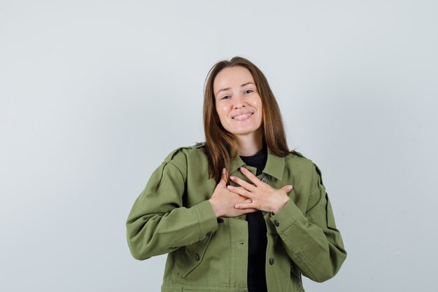 Mujer joven cogidos de la mano en el pecho con chaqueta verde y mirando agradecido. vista frontal.