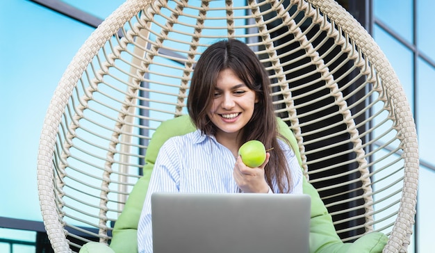 Una mujer joven come una manzana y trabaja en una computadora portátil.