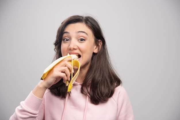 Mujer joven comiendo plátano sobre pared gris.