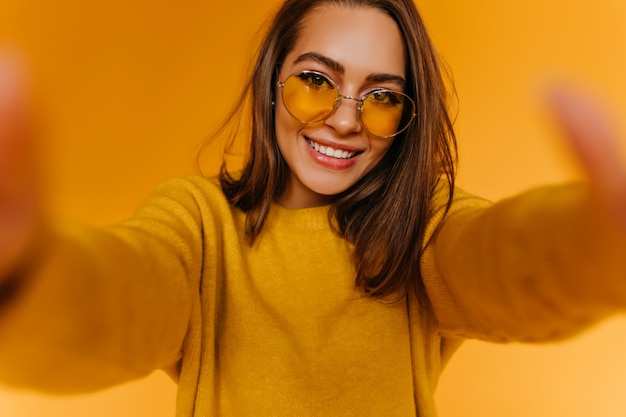 Mujer joven complacida con cabello castaño haciendo selfie en pared amarilla con sonrisa encantadora. Chica atractiva tomando una foto de sí misma y riendo.