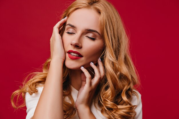 Mujer joven complacida con cabello rubio posando con los ojos cerrados. Chica rubia despreocupada de pie en la pared roja.