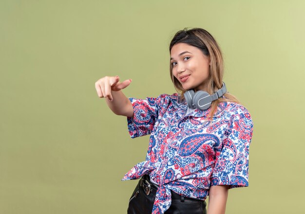 Una mujer joven complacida en camisa estampada de paisley usando audífonos apuntando con el dedo índice mientras mira el en una pared verde