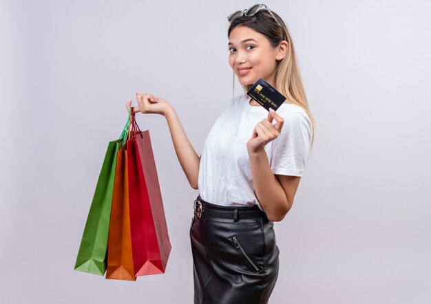 Una mujer joven complacida en camiseta blanca con gafas de sol en la cabeza mostrando la tarjeta de crédito mientras sostiene bolsas de la compra en una pared blanca