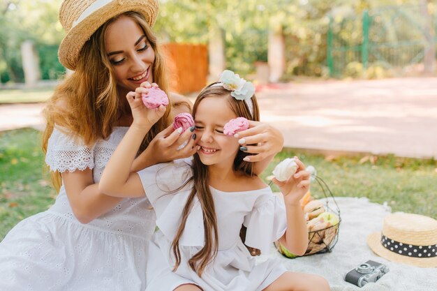 Mujer joven complacida y niña emocionada en atuendos blancos sentados en una manta con canasta de frutas. Foto al aire libre de dama elegante jugando con su hija durante el almuerzo.