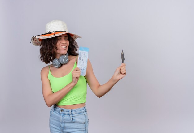 Una mujer joven complacida con el pelo corto en la parte superior verde de la cosecha en auriculares con sombrero para el sol mirando boletos de avión y tarjeta de crédito sobre un fondo blanco.