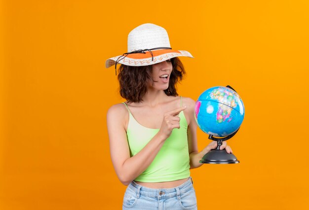 Una mujer joven complacida con el pelo corto en verde crop top con sombrero para el sol apuntando a un globo con el dedo índice