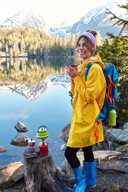 Mujer joven complacida sonríe suavemente, bebe bebidas calientes, usa impermeable y botas de goma, disfruta del clima soleado después de la lluvia