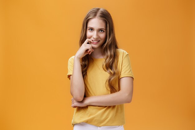 Mujer joven complicada creativa con cabello largo ondulado natural en camiseta amarilla mirando desde debajo de la frente con intención y lujuria en expresión mordiendo el dedo, sonriendo a la cámara sobre la pared naranja.