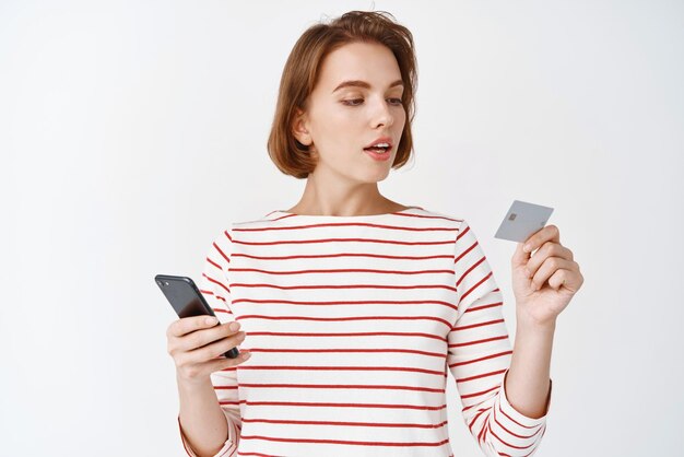 Mujer joven comprando en línea mirando una tarjeta de crédito de plástico para pagar en un teléfono móvil de pie contra el fondo blanco