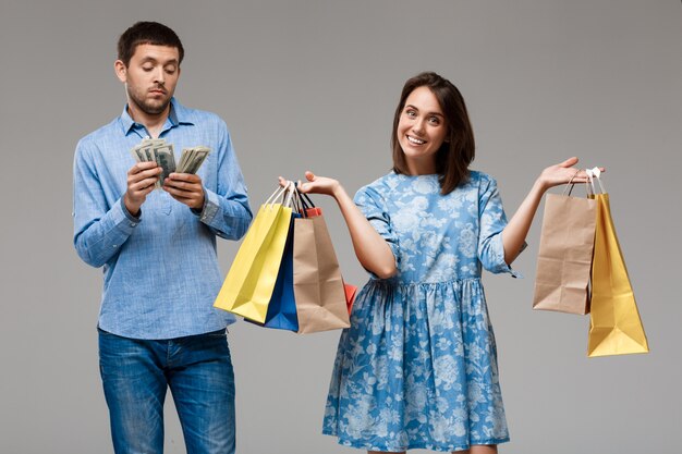 Mujer joven con compras y hombre con dinero sobre pared gris