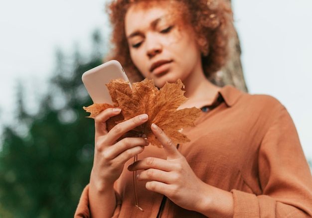 Foto gratuita mujer joven comprobando su teléfono mientras sostiene una hoja seca