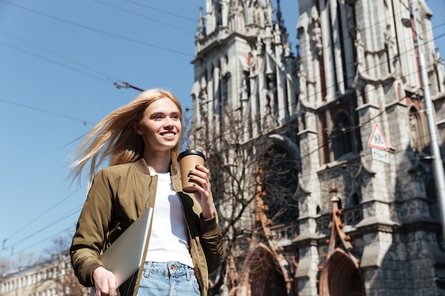 Mujer joven con computadora portátil y café caminando en la calle