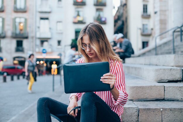 Una mujer joven con una computadora portátil sentada en las escaleras, cerca de la universidad