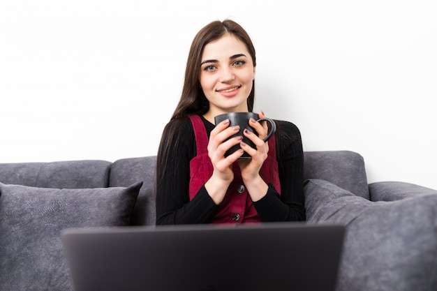 Mujer joven con la computadora portátil en el sofá. Una niña pasa mucho tiempo en Internet en su computadora portátil. Mujer relajante en la casa después de un baño con una computadora portátil y una taza de té.