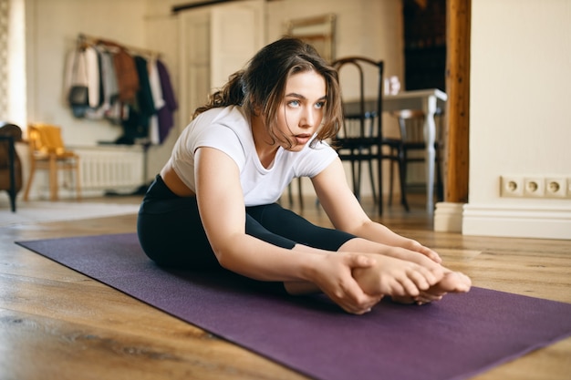 Mujer joven concentrada con cuerpo flexible sentada en una colchoneta con las piernas estiradas, haciendo flexión hacia adelante sentada o Paschimottanasana, inclinada sobre las rodillas, manteniendo los dedos de los pies en punta.