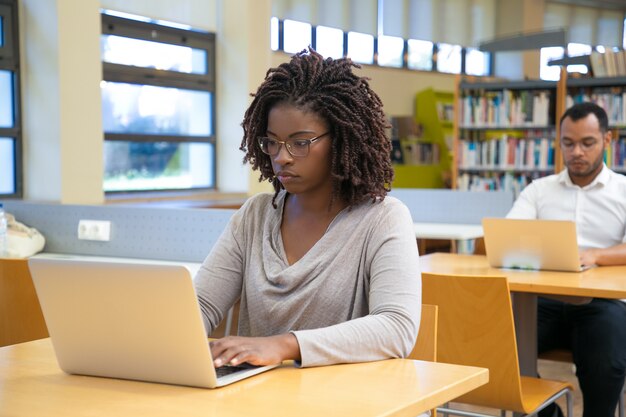 Mujer joven concentrada que trabaja con la computadora portátil en la biblioteca