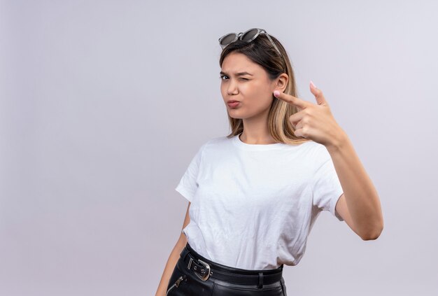 Una mujer joven confiada en camiseta blanca con gafas de sol en la cabeza apuntando a algo con el dedo índice mientras mira en una pared blanca