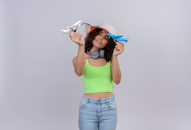 Una mujer joven confiada con el pelo corto en la parte superior verde de la cosecha en auriculares con sombrero para el sol sosteniendo aviones de juguete azul y blanco en las manos sobre un fondo blanco.