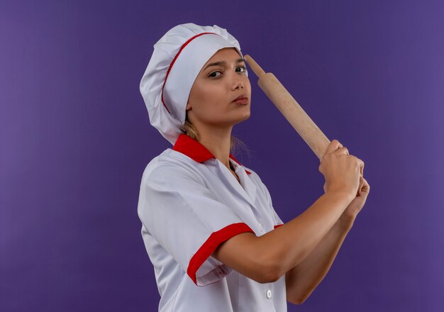 Mujer joven confidente del cocinero que lleva uniforme del cocinero que sostiene el rodillo en la pared aislada con el espacio de la copia