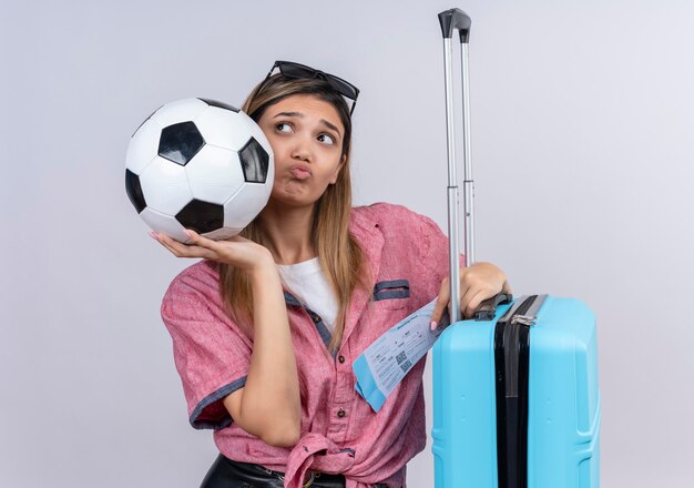 Una mujer joven confundida con camisa roja y gafas de sol mirando hacia la cámara mientras sostiene la bola con boletos de avión y maleta azul en una pared blanca