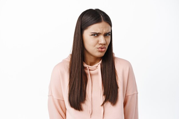 Mujer joven confundida frunciendo el ceño y haciendo muecas, encogerse de algo extraño y confuso, de pie sobre una pared blanca