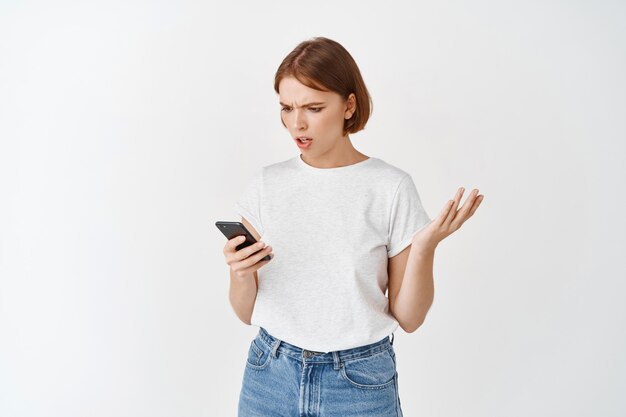 Mujer joven confundida mirando el teléfono móvil con expresión cuestionada, encogiéndose de hombros preocupada, de pie contra la pared blanca