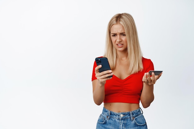 Mujer joven confundida mirando el teléfono móvil mientras tiene problemas con la tarjeta de crédito con el pedido o el pago sobre fondo blanco