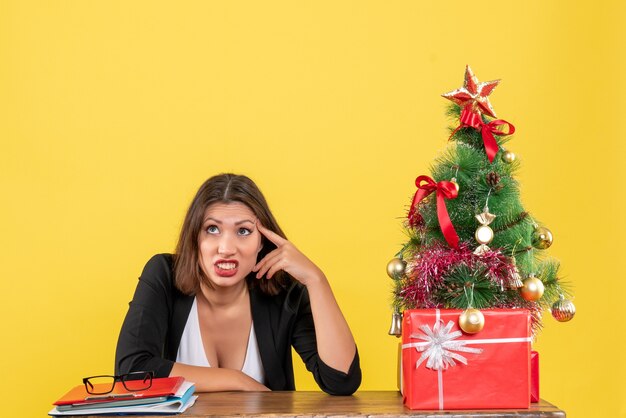 Mujer joven confundida y nerviosa sentada en una mesa cerca del árbol de Navidad decorado en la oficina en amarillo