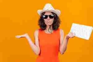 Foto gratuita una mujer joven confundida con el pelo corto en una camisa naranja con sombrero para el sol y gafas de sol levantando las manos con el mapa sin saber qué hacer