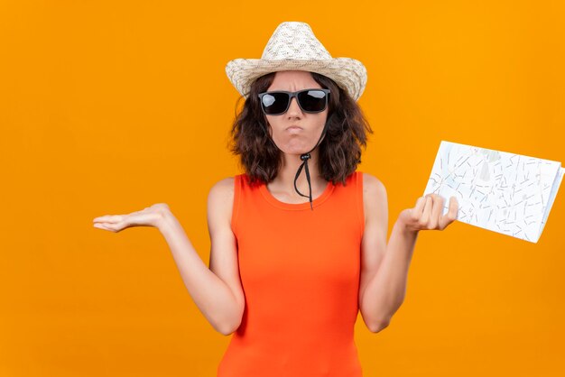 Una mujer joven confundida con el pelo corto en una camisa naranja con sombrero para el sol y gafas de sol levantando las manos con el mapa sin saber qué hacer