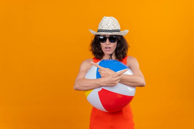 Una mujer joven confundida con el pelo corto en una camisa naranja con sombrero para el sol y gafas de sol sosteniendo una bola colorida inflable apuntando a algo