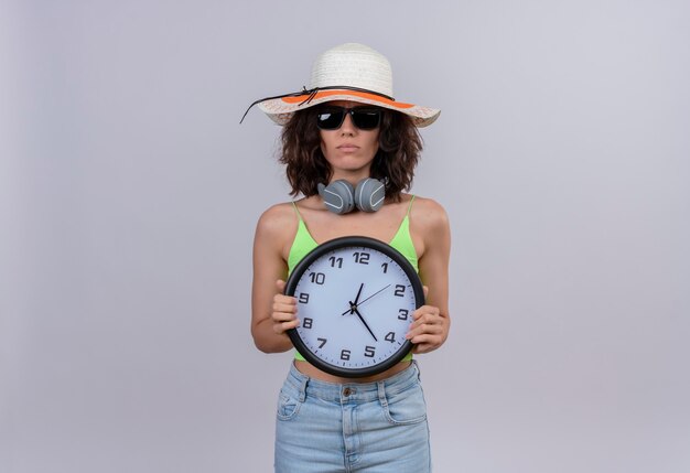 Una mujer joven confundida con el pelo corto en la parte superior verde con gafas de sol y sombrero para el sol sosteniendo un reloj de pared sobre un fondo blanco.