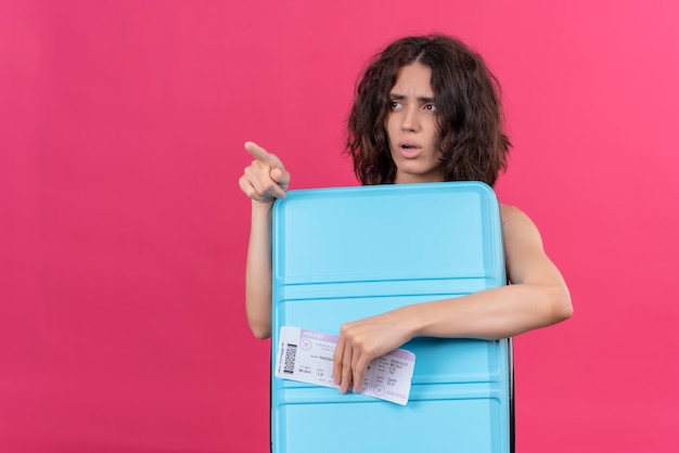 Una mujer joven confundida con pelo corto vistiendo verde crop top apuntando con el dedo índice sosteniendo la maleta azul con billetes de avión