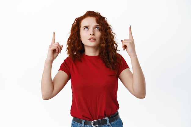 Mujer joven confundida con el pelo rizado rojo que tiene dudas apuntando con el dedo hacia arriba y mirando hacia arriba con la cara cuestionada ve algo extraño fondo blanco