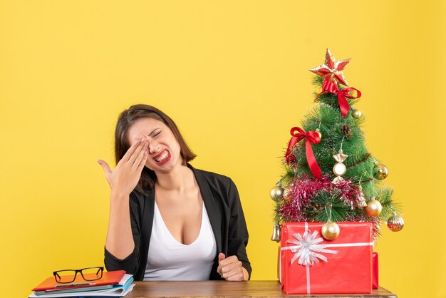 Mujer joven confundida sentada en una mesa en traje cerca del árbol de Navidad decorado en la oficina en amarillo