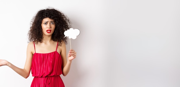 Foto gratuita mujer joven confundida con vestido rojo frunciendo el ceño y mirando molesta sosteniendo un palo de nube de comentarios de pie ag