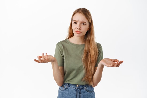 Mujer joven confusa y seria encogiéndose de hombros, levantando las manos hacia los lados perplejo, no sé nada, no puedo decir, mira fijamente interrogada al frente, de pie sobre una pared blanca