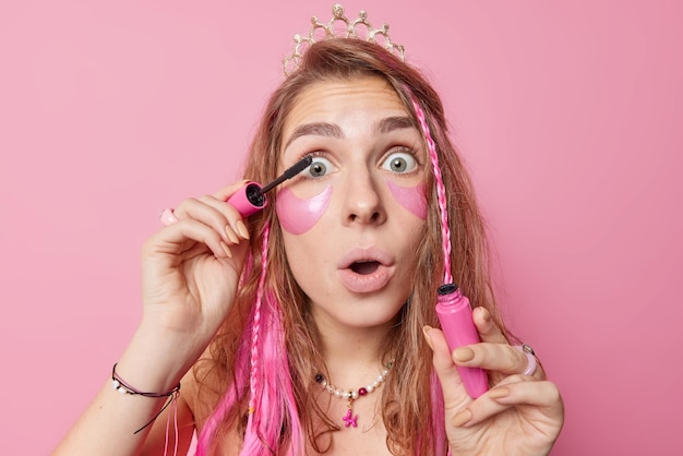 Mujer joven conmocionada con cabello largo que aplica rímel tiene los ojos muy abiertos y la boca usa parches de hidrogel usa corona quiere tener una apariencia fabulosa en la fiesta aislada sobre el fondo rosado del estudio