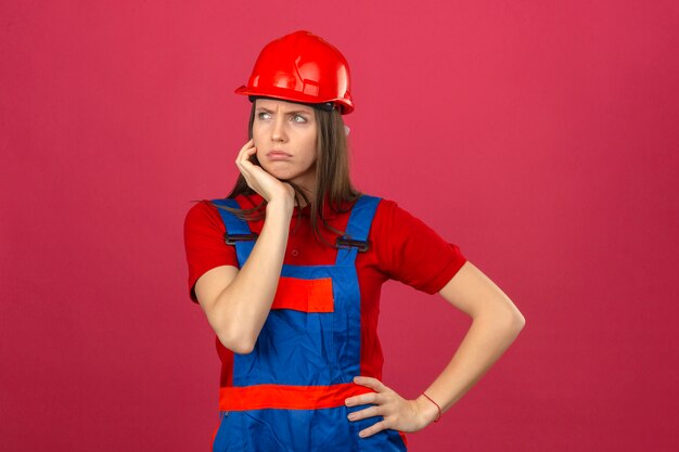 Mujer joven en construcción uniforme y casco de seguridad rojo mirando hacia los lados con expresión dudosa y escéptica sobre fondo rosa oscuro