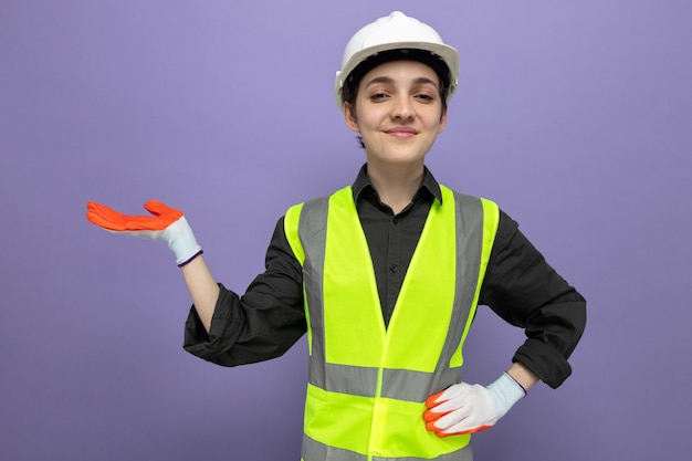 Mujer joven constructor en chaleco de construcción y casco de seguridad en guantes de goma sonriendo confiado presentando espacio de copia con el brazo de su mano de pie en azul