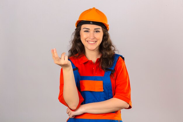 Mujer joven constructor en construcción uniforme y casco de seguridad sonriendo presentando e invitando a venir con la mano sobre la pared blanca aislada