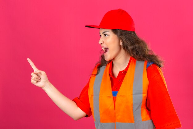 Mujer joven constructor en uniforme de construcción y casco de seguridad acusando a alguien sobre pared rosa aislada